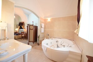 Une salle de bain avec une grande baignoire, un lavabo et un meuble-lavabo. Il y a un miroir au-dessus du lavabo et un porte-serviettes à côté. La pièce est dotée d’un sol carrelé et d’une fenêtre avec des rideaux.