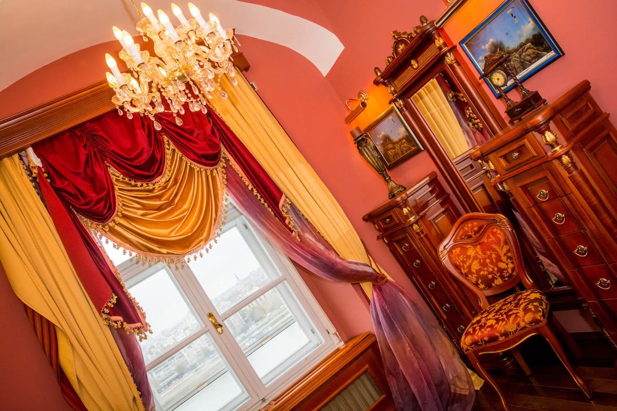 A room with a large window, ornate curtains, a chandelier, a wooden cabinet, and a chair.
