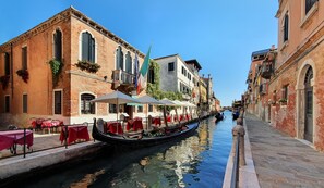 A narrow canal with a gondola, lined with buildings featuring outdoor seating and flags. - Messner Palace (Venice)