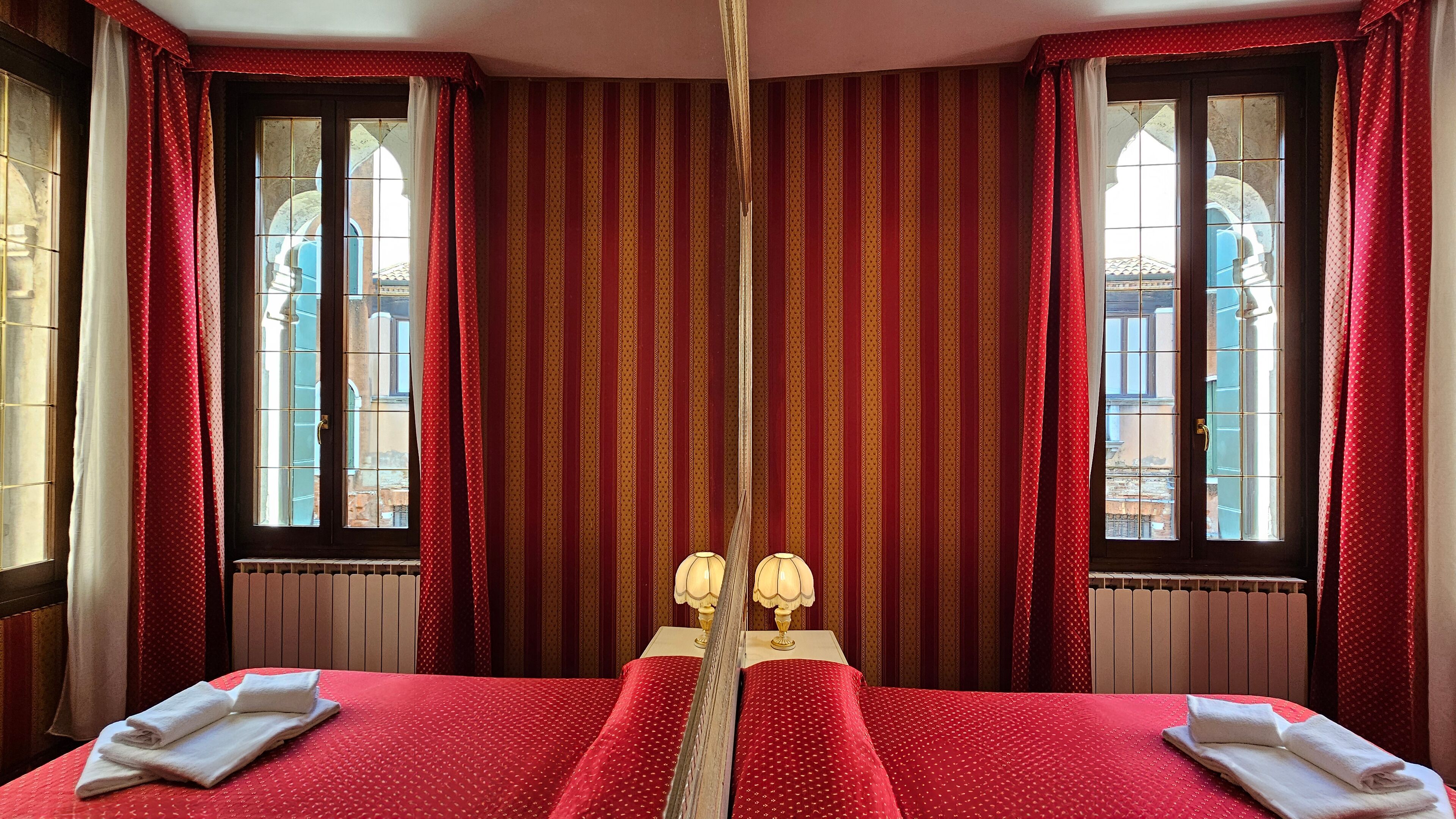 Habitación de hotel con una cama grande, dos lámparas de noche y vistas a edificios a través de las ventanas.