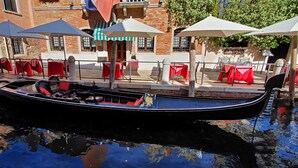Un canal con una góndola, mesas con manteles rojos y sombrillas.