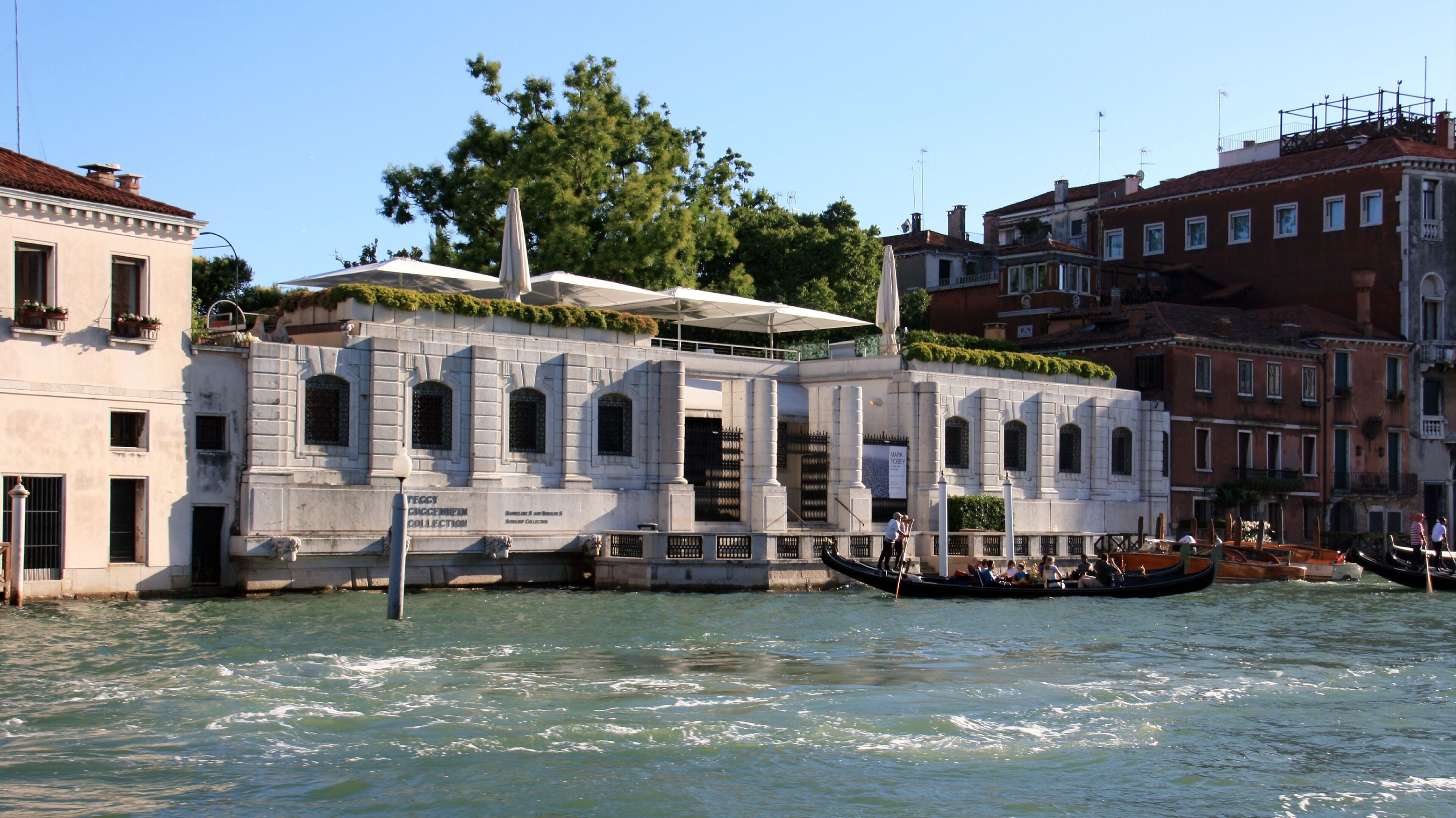 A historic building with a stone facade, a covered area with plants, and a canal with gondolas.