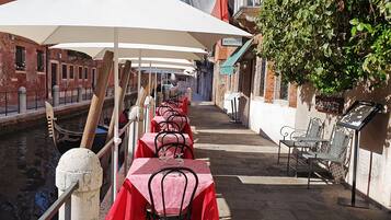 Un restaurant situé au bord d’un canal, avec des tables protégées par des parasols blancs, des nappes rouges et des chaises en métal.