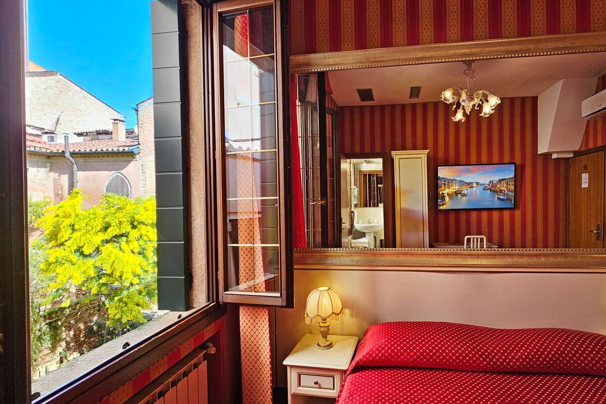 Habitación de hotel con un ventanal grande, una cama con ropa de cama roja, dos lámparas de noche, un espejo y un televisor que muestra un paisaje urbano.