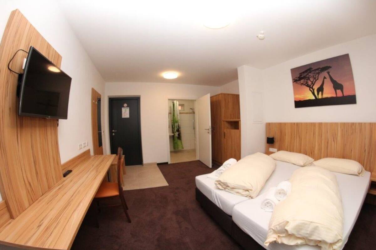 A hotel room with a bed, a desk, a chair, a TV mounted on the wall, and a painting of giraffes.