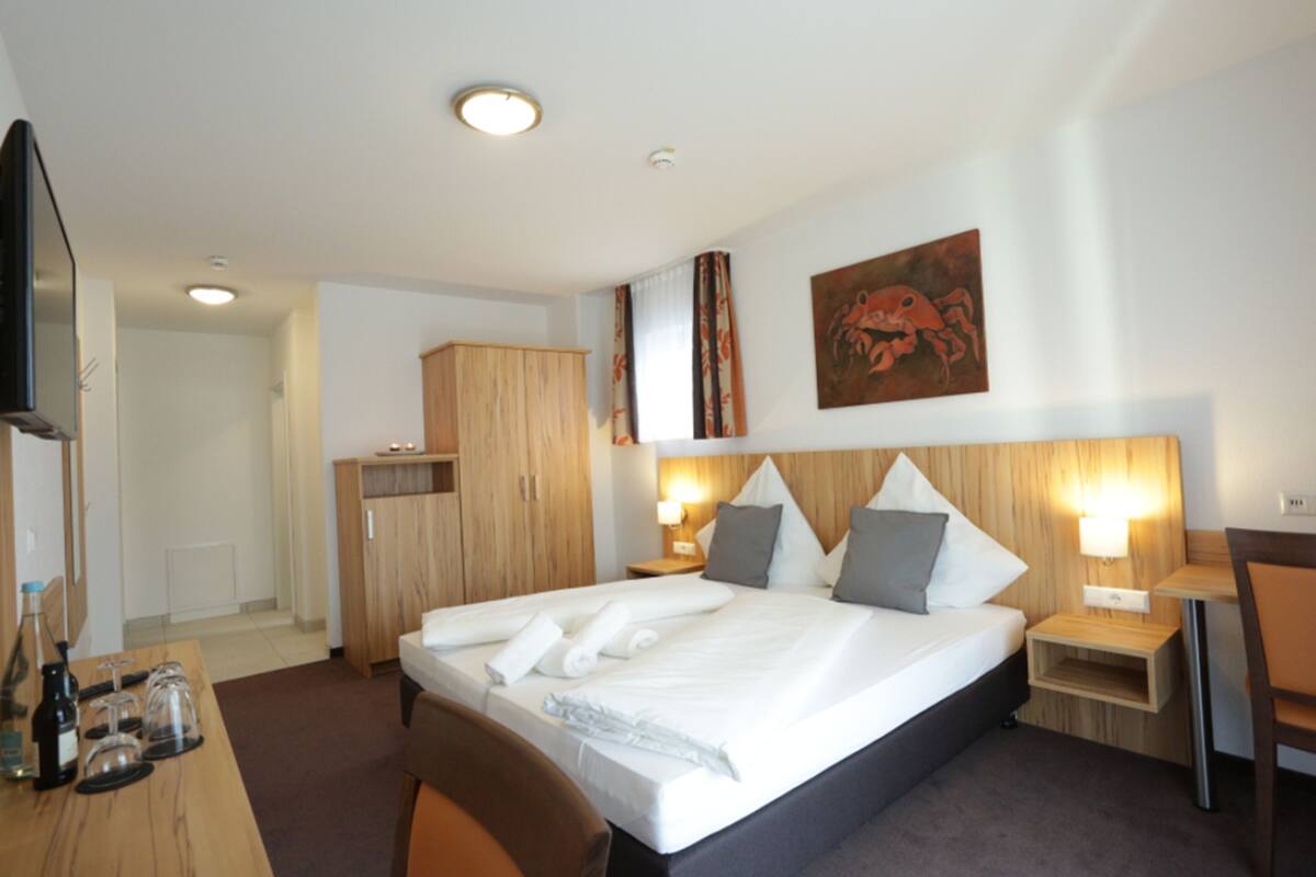 A hotel room with a large bed, a wooden wardrobe, a desk with a chair, and a painting on the wall.