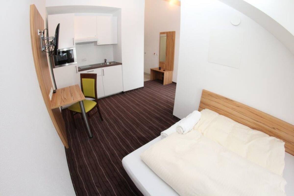 A compact hotel room with a bed, a small desk, a chair, a kitchenette, and a mirror.