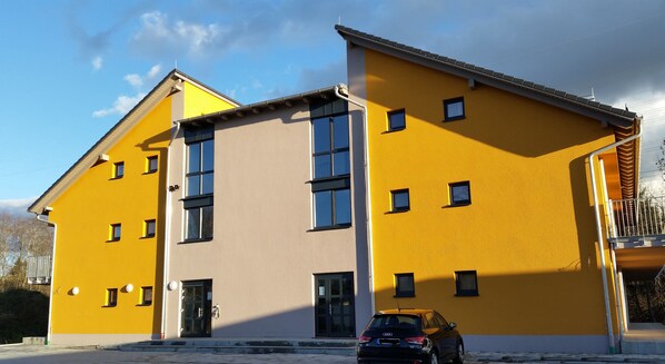 Ein zweistöckiges Gebäude mit gelber Fassade, davor ein schwarzes Auto und ein klarer Himmel.