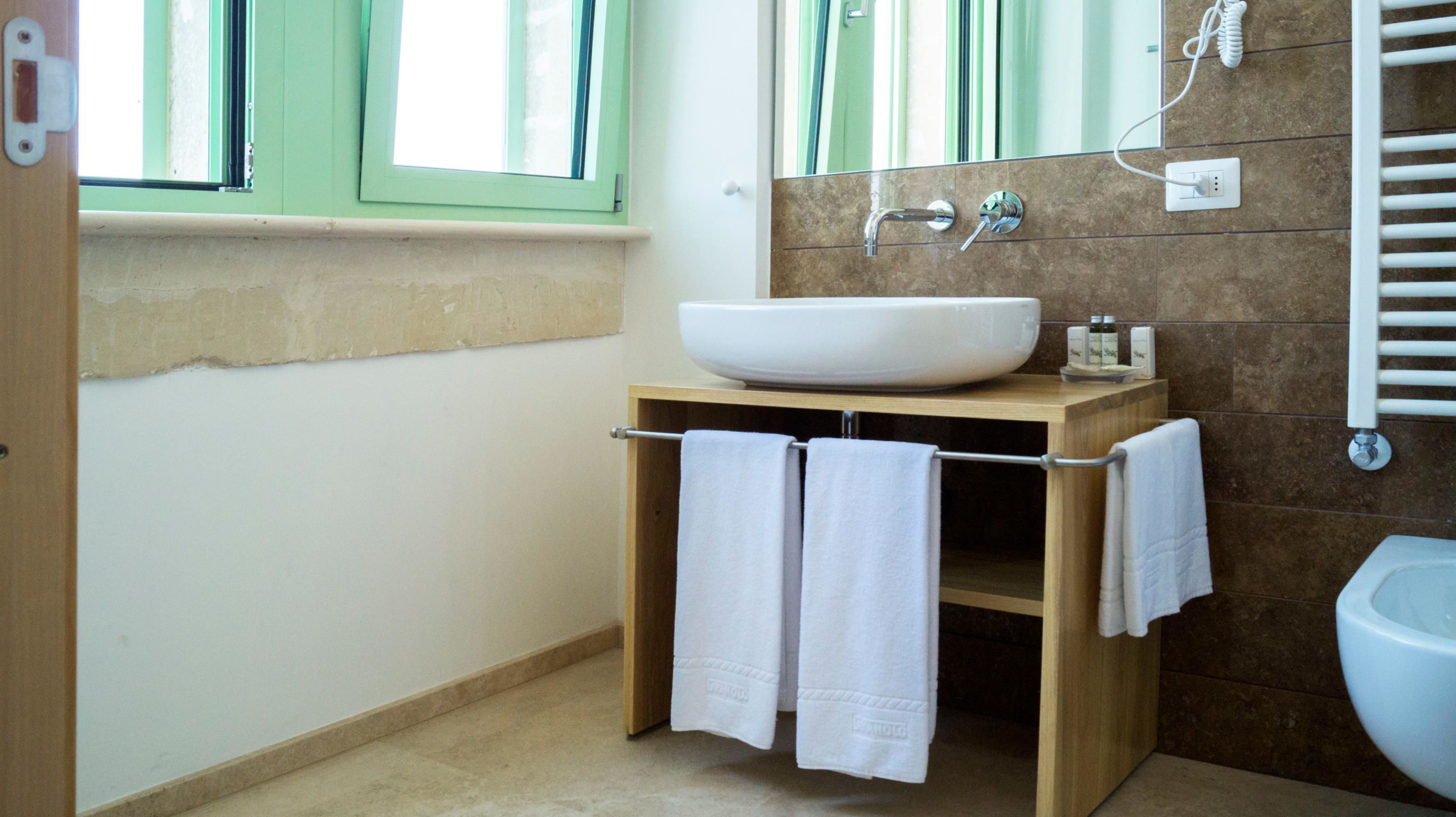 A bathroom with a large white sink, wooden cabinet, two towels hanging, a window with green shutters, and a white toilet.