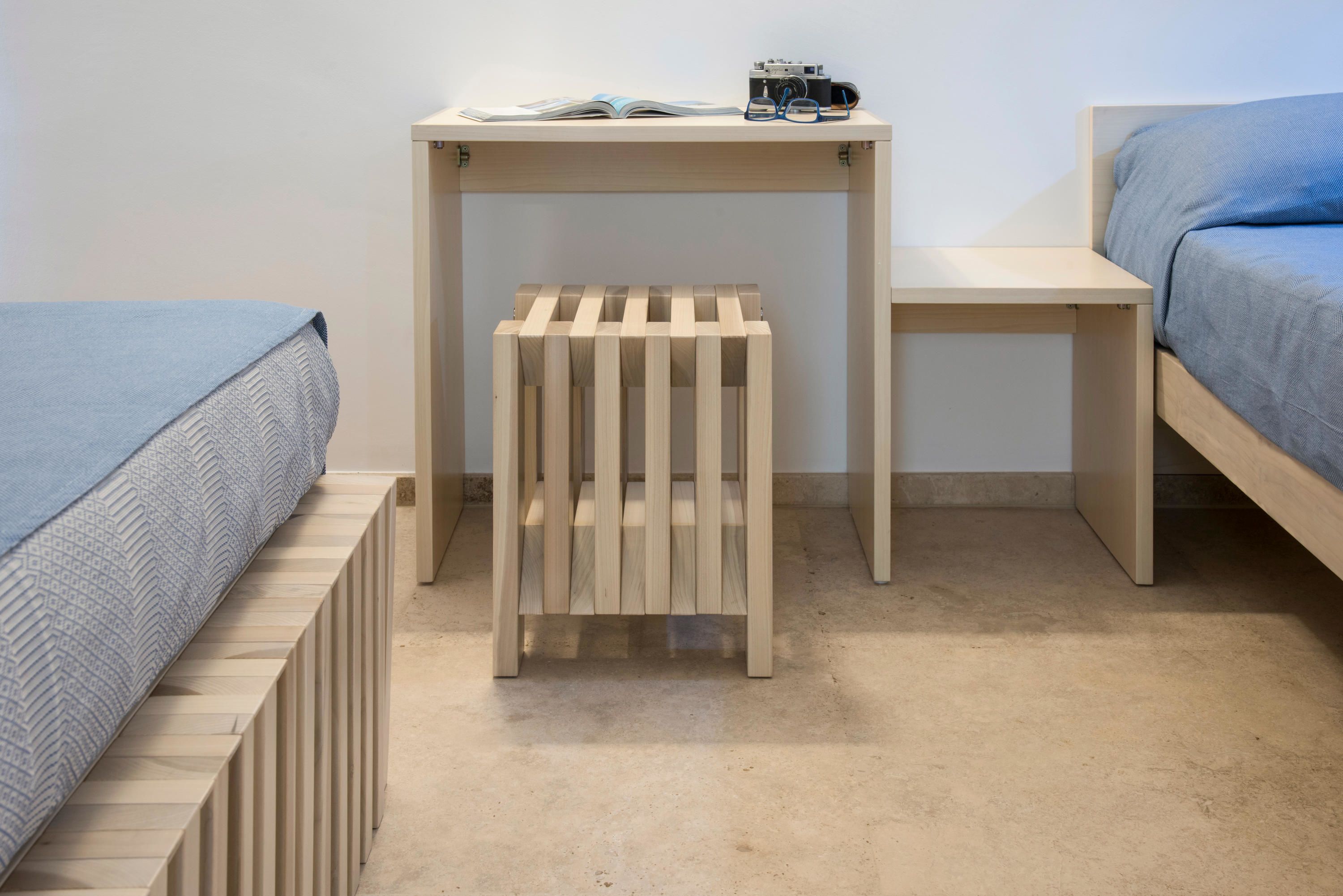 A bedroom with a bed, a small wooden stool, a nightstand, and a desk with a book and glasses on it.