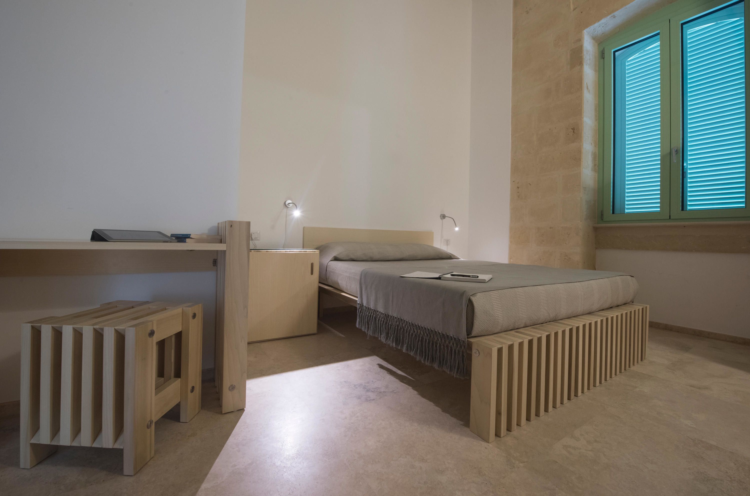 A minimalist bedroom with a bed, bedside tables, a small wooden stool, and a window with blinds.