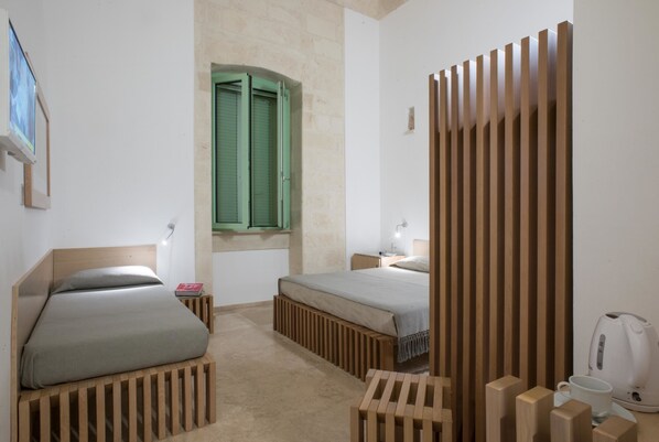 Un dormitorio con dos camas, una puerta verde, un separador de madera, una mesita de noche con lámpara y un hervidor en la mesita auxiliar.