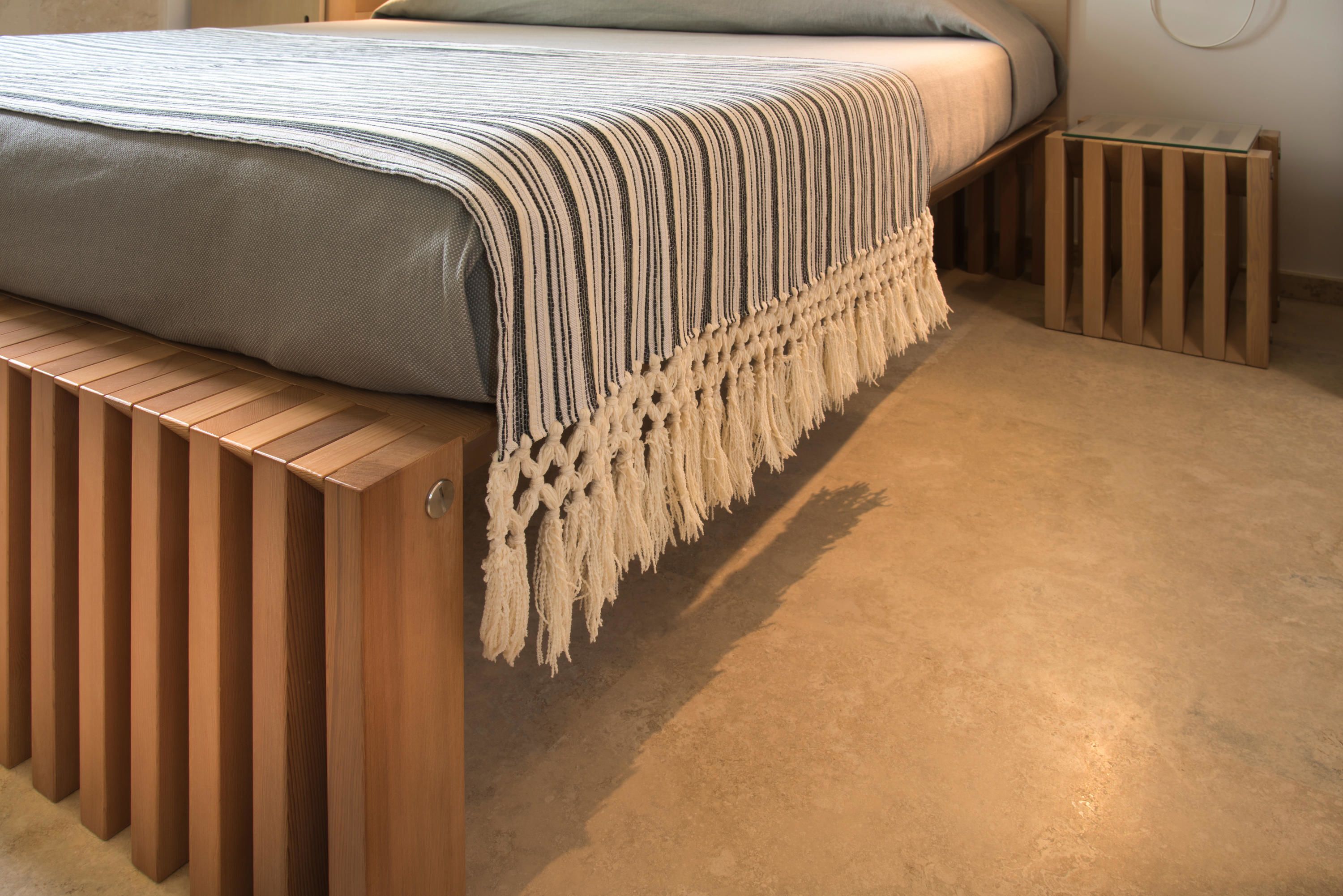 Una cama con un edredón a rayas y un chal de macramé, un banco de madera y una mesita con superficie de vidrio.
