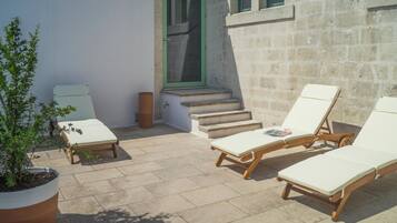A sunlit outdoor seating area with two lounge chairs and a small table, a potted plant, and a stone wall.