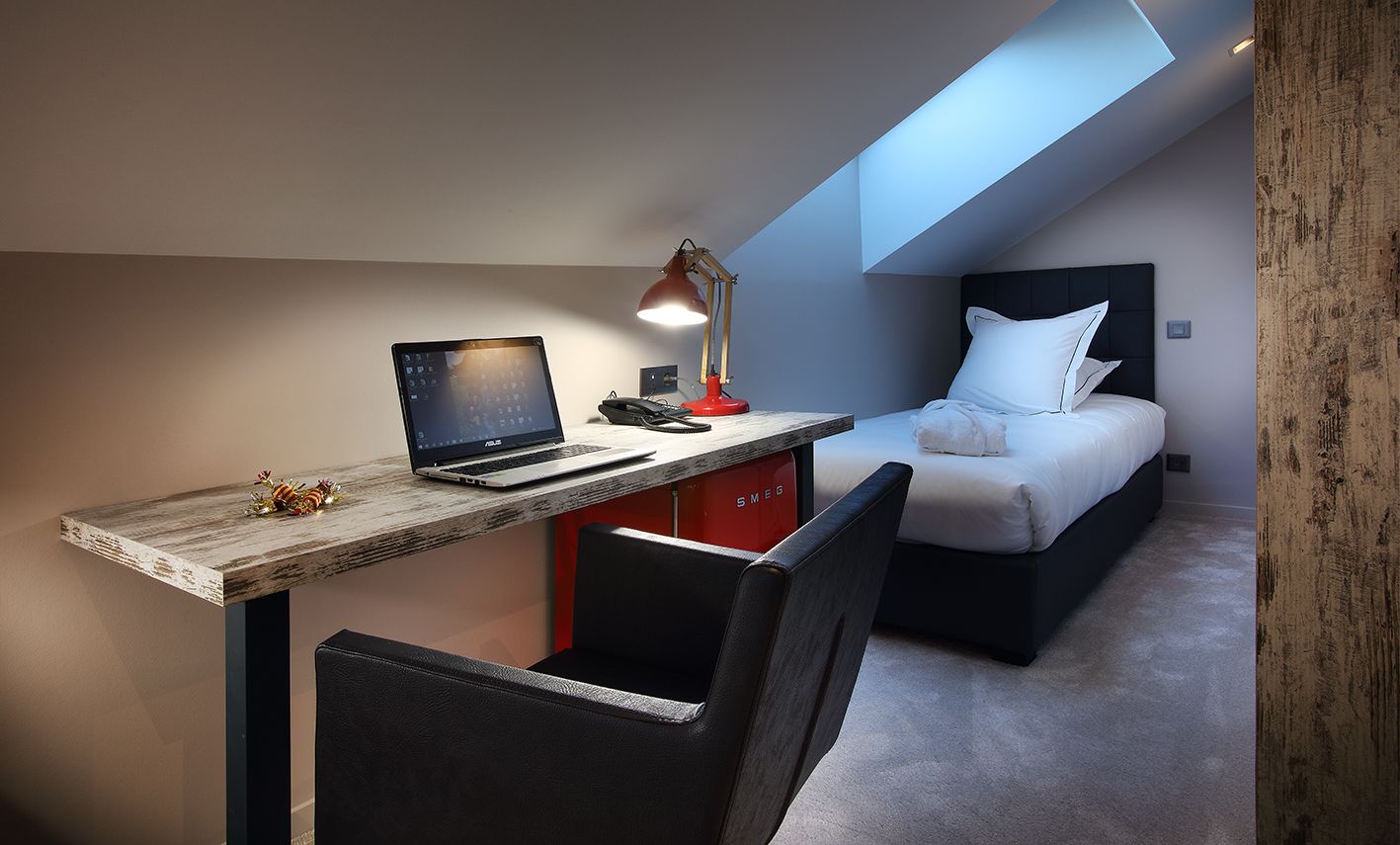 Une chambre d’hôtel compacte, équipée d’un bureau, d’un ordinateur portable et d’un lit avec une literie blanche.
