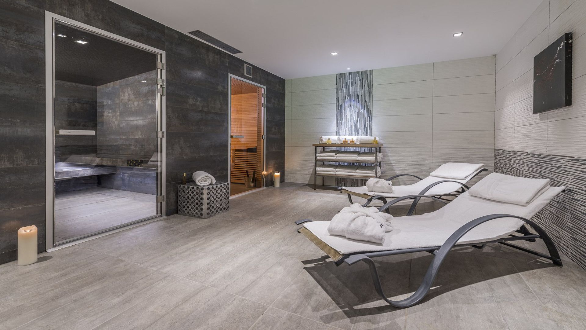 A modern spa room with a glass shower enclosure, a wooden bench, a massage table, and a shelf with towels and toiletries.