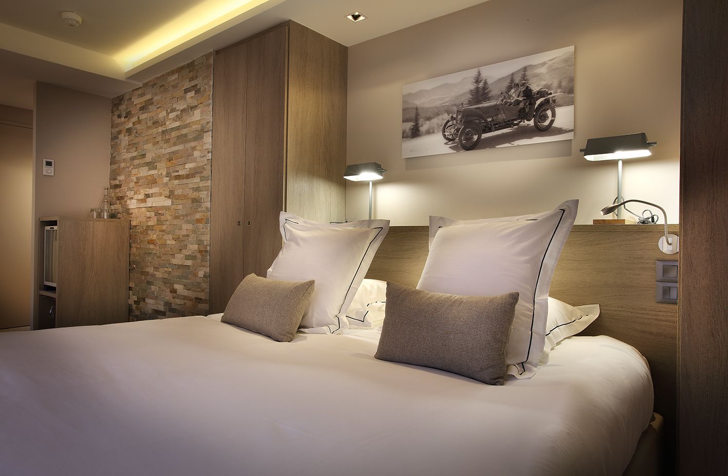 Ein Hotelzimmer mit einem großen Bett, zwei Nachttischlampen, einer Wand mit Steinverkleidung und einem Vintage-Automobilbild.