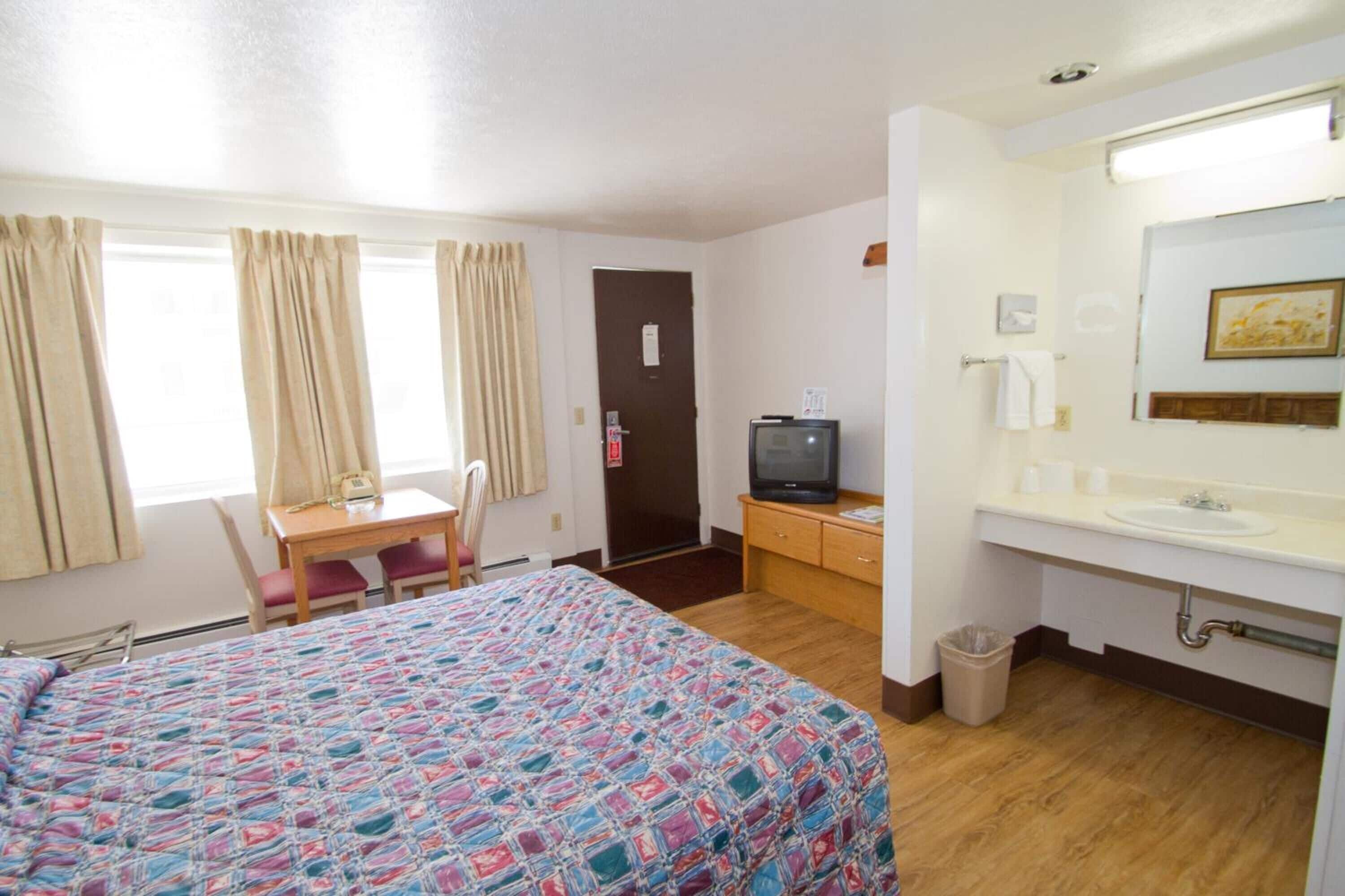 Une chambre d’hôtel comprenant un lit, un bureau avec une chaise, une télévision, une salle de bain avec un lavabo et un miroir, et une poubelle.
