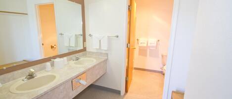 Une salle de bain avec deux lavabos, un grand miroir et une porte donnant sur un espace où se trouvent les toilettes.