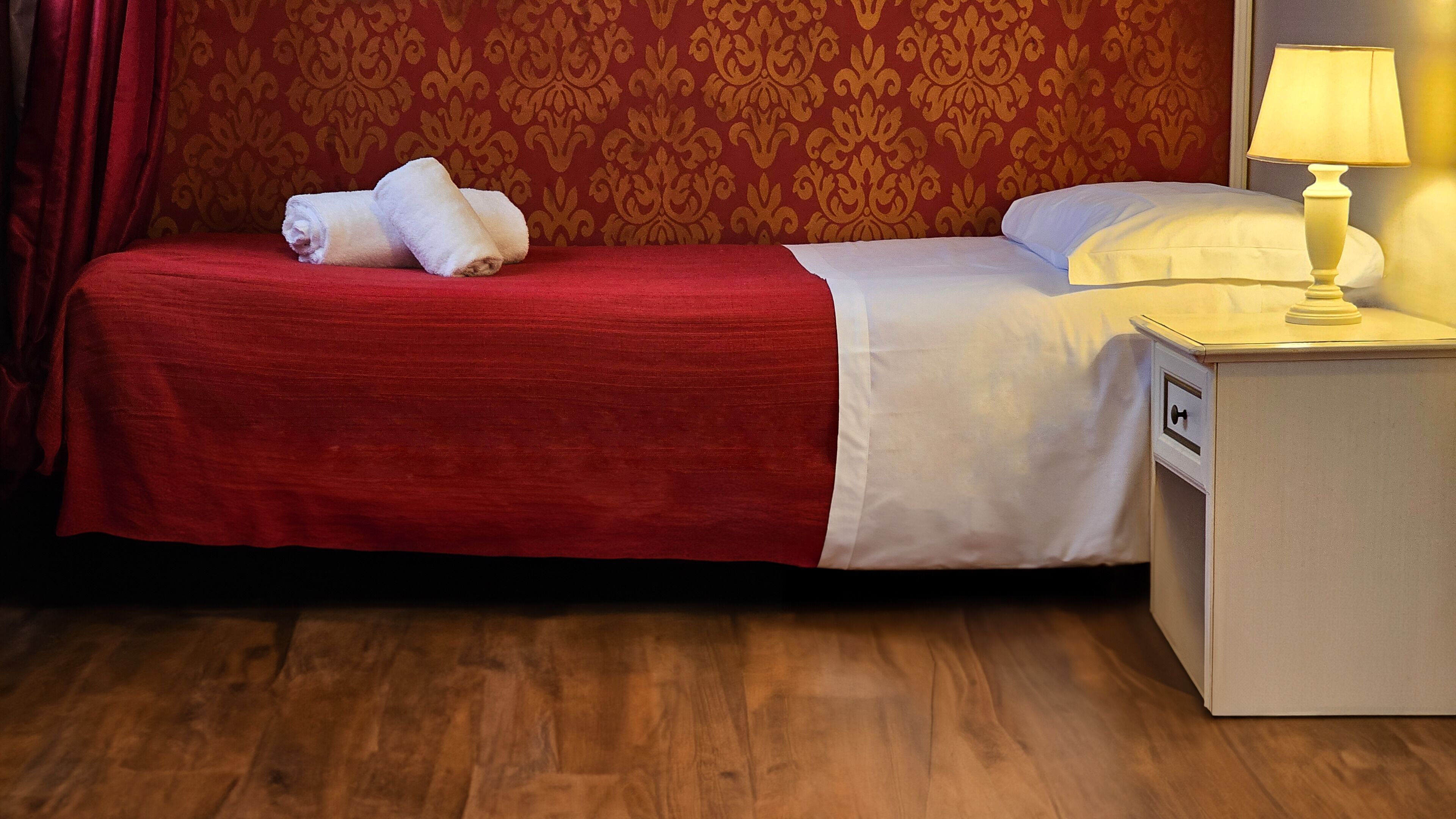 A neatly made bed with a red bedspread and white pillows, a white nightstand with a lamp, and a wooden floor.