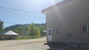 A single-story building with a flat roof, a door, and a small window. There is a covered area with a roof to the left. The building is surrounded by a paved area with a fence and trees in the background. - Sleep Right Inn (Enderby)