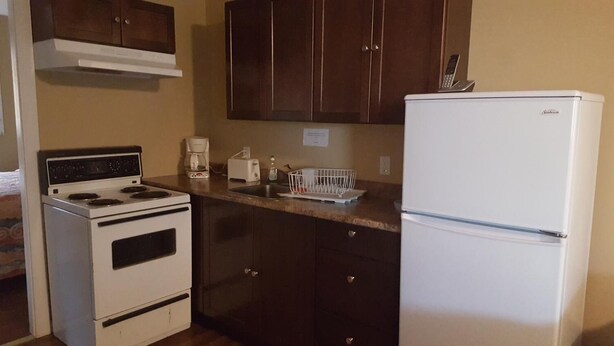 A compact kitchen with a stove, oven, and refrigerator, wooden cabinets, and a countertop with a coffee maker and toaster. - Sleep Right Inn (Enderby)