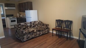 A living room with a patterned sofa, two wooden chairs, a television, a kitchen area with a refrigerator, stove, and cabinets.