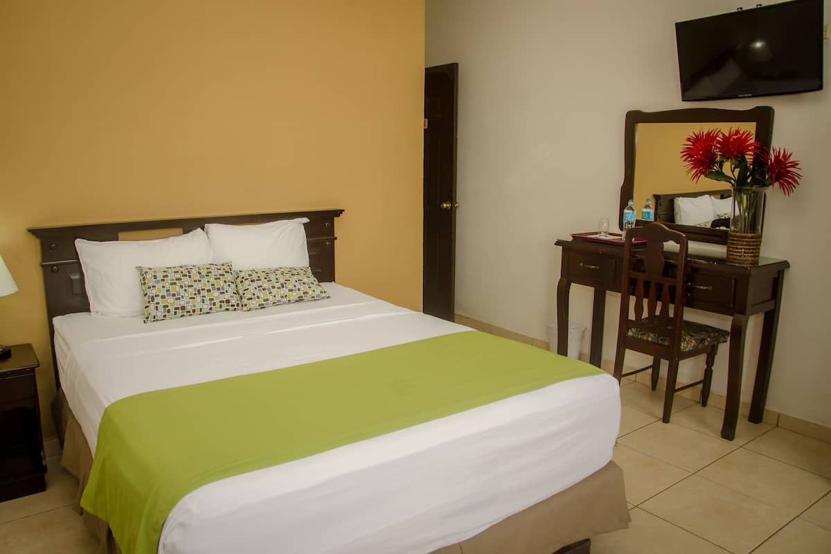 A neatly made bed with a green bedspread, a wooden nightstand with a lamp, a desk with a chair, and a mirror on the wall.