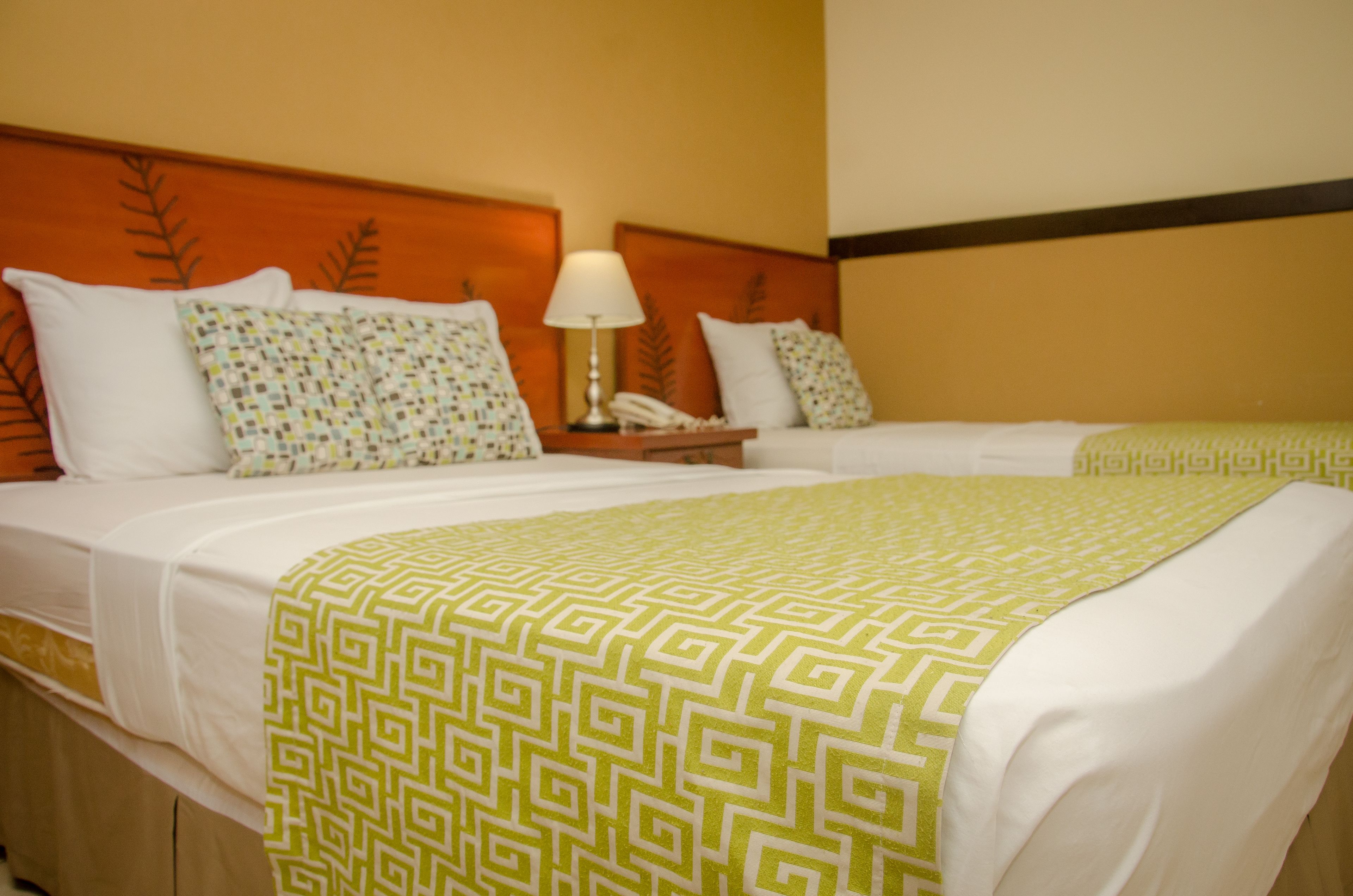 A hotel room with two beds, each with white linens and patterned pillows. There is a nightstand with a lamp and a phone between the beds.