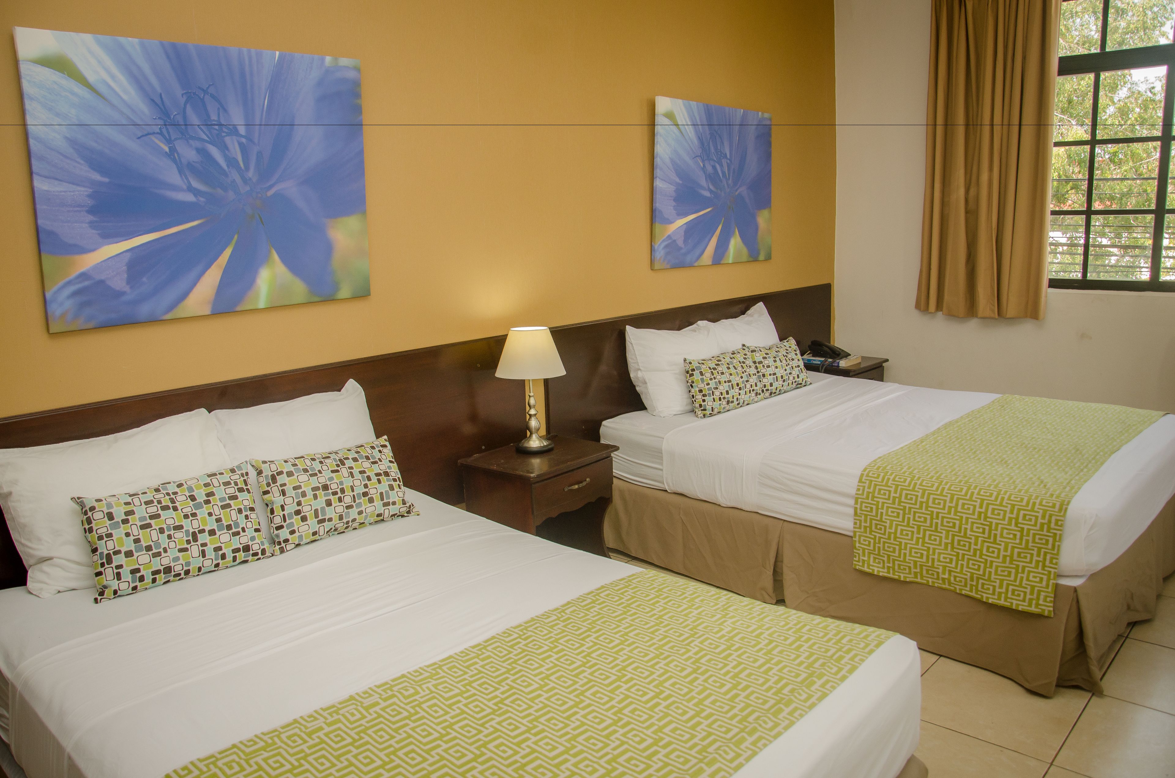 A hotel room with two beds, each with a patterned pillow and a green throw. There are two wall-mounted lamps and two paintings of blue flowers.