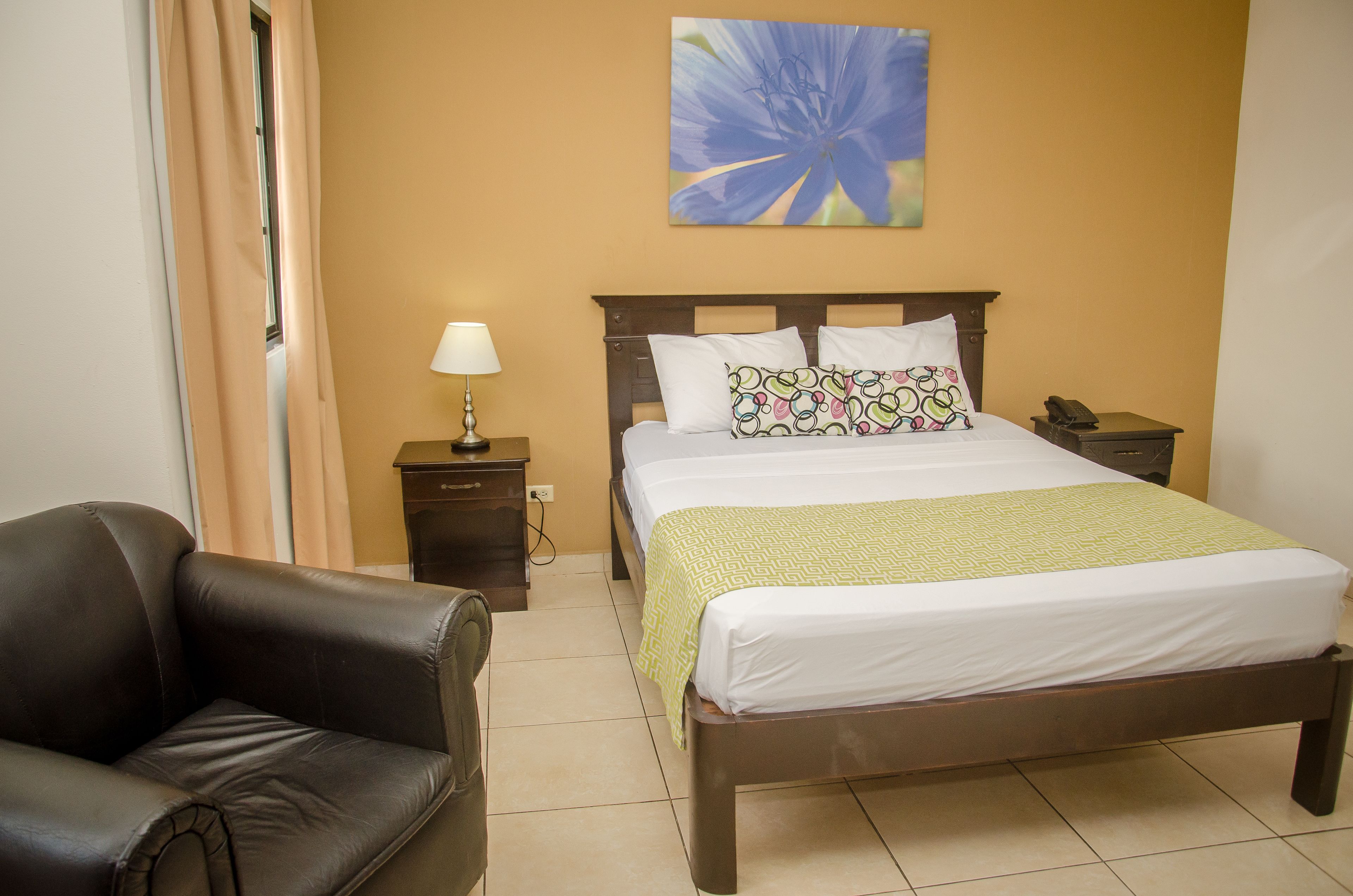 A neatly arranged hotel room with a bed, a chair, a bedside table with a lamp, and a wall-mounted painting.