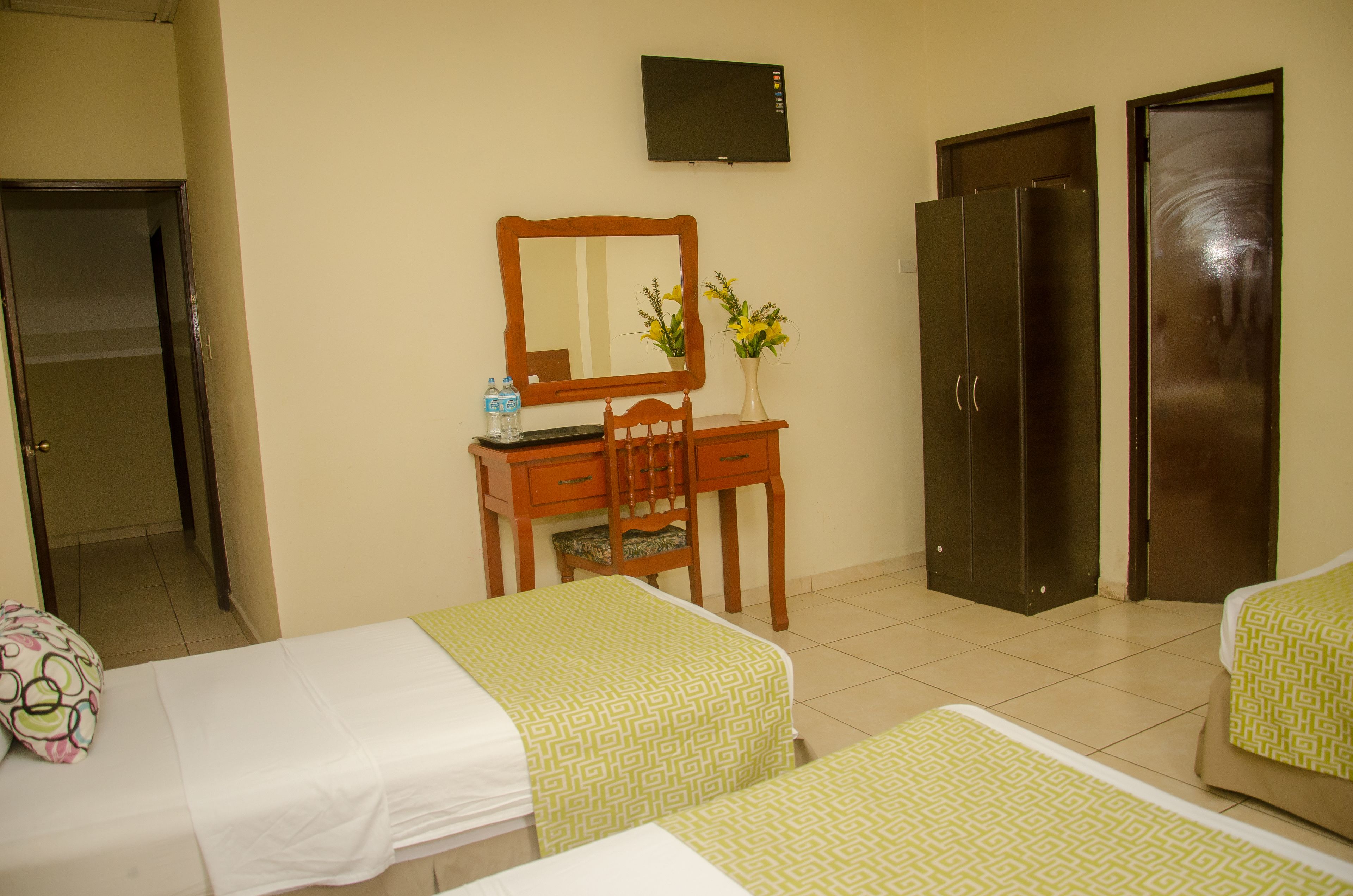 Habitación de hotel con dos camas, un escritorio con silla, un espejo, un televisor y dos puertas.