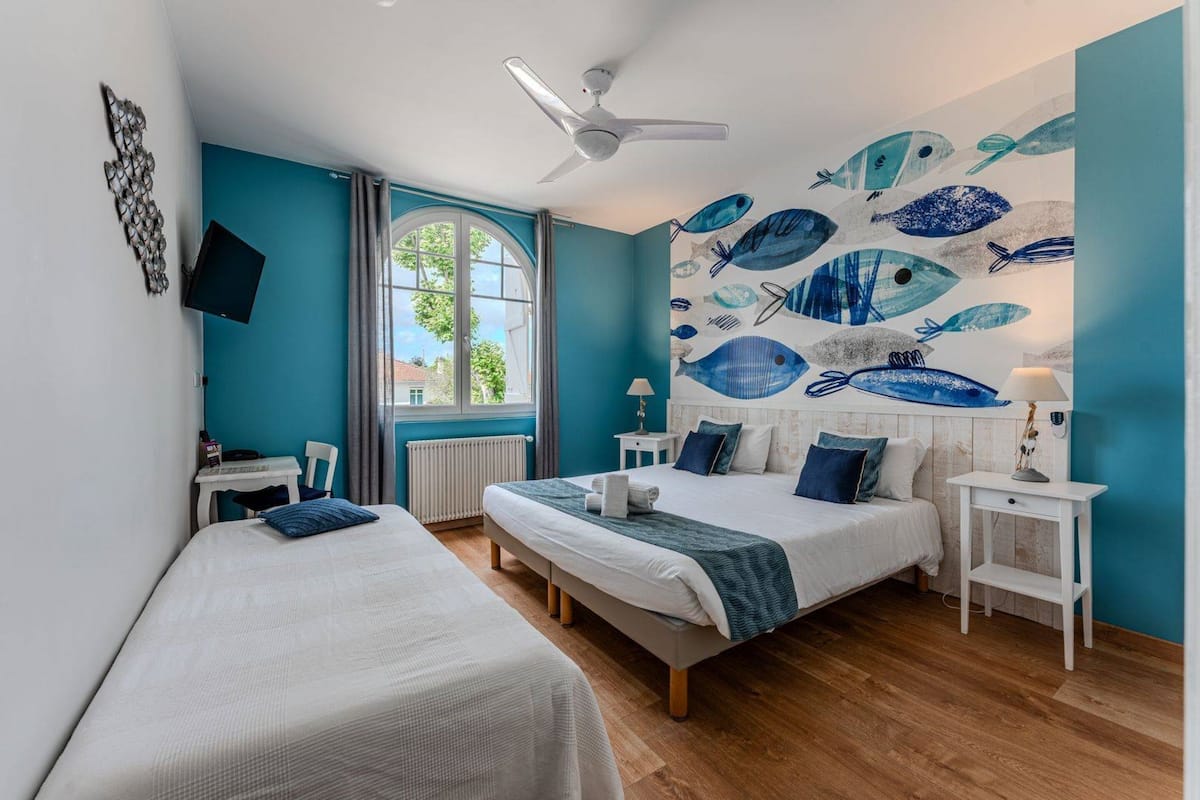 A hotel room with two beds, a ceiling fan, a large window with curtains, a wall mural of fish, and a TV mounted on the wall.