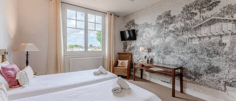 A hotel room with two beds, a desk, a chair, a ceiling fan, a TV, and a large window with curtains.