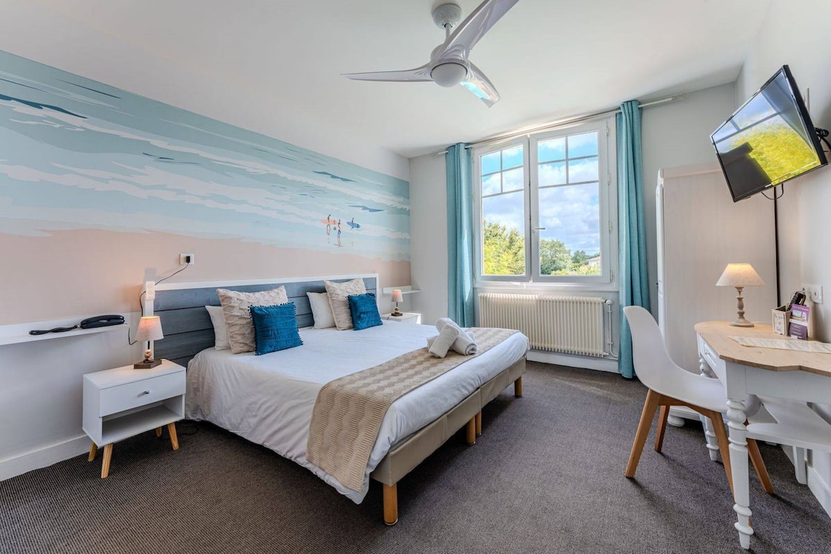 A bedroom with a bed, bedside table, desk, chair, ceiling fan, window, and a TV.