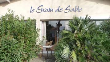 El exterior de 'le Grain de Sable', con una entrada de puerta de vidrio, rodeado de exuberante follaje y un pequeño área de asientos al aire libre.