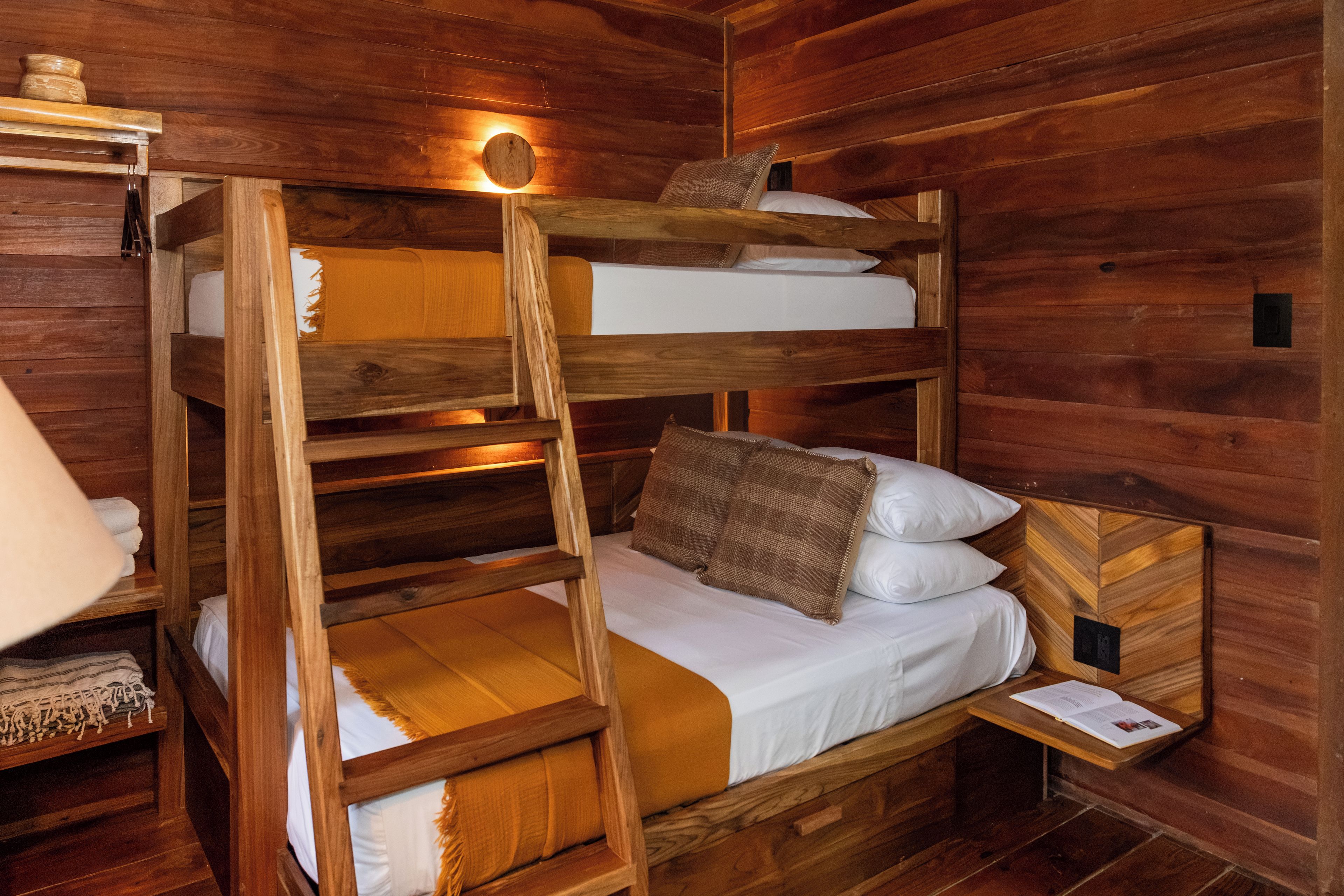 A bunk bed with a ladder, wooden walls, a small bedside table with a lamp, and a shelf with towels.