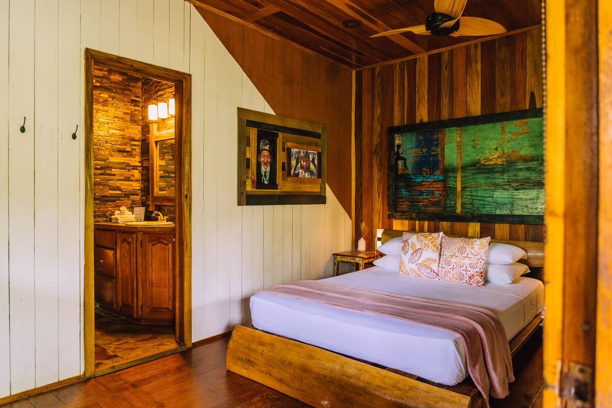 A bedroom with a wooden headboard, a bed with a white and pink bedspread, a nightstand with a lamp, and a painting on the wall.
