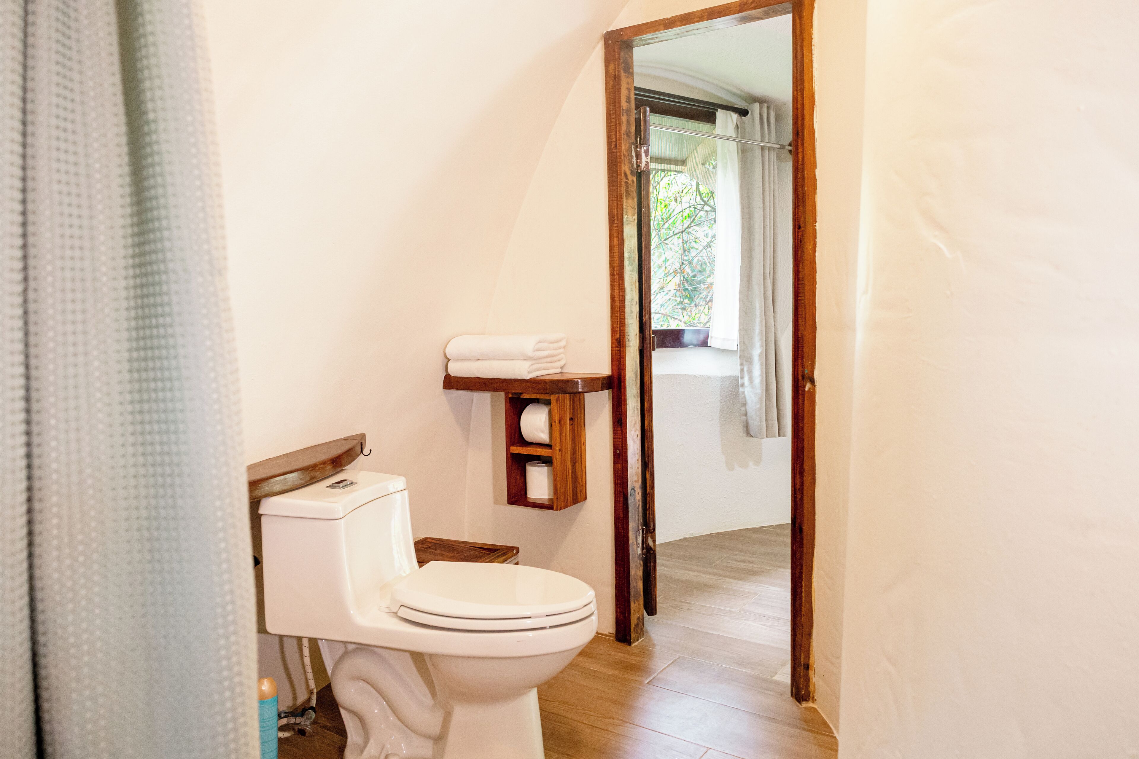 A bathroom with a toilet, a wooden shelf with towels, and a view through an open door to a balcony.
