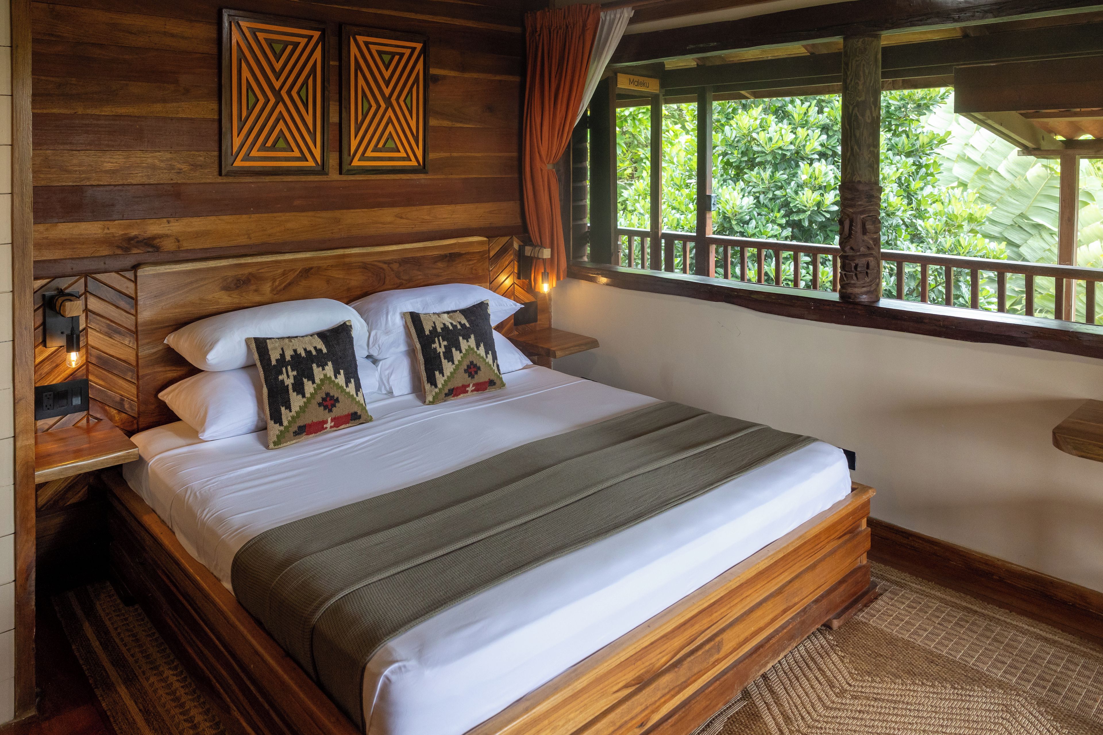 A wooden-framed bed with white linens and patterned pillows, a nightstand with a lamp, and a window with a view of greenery.