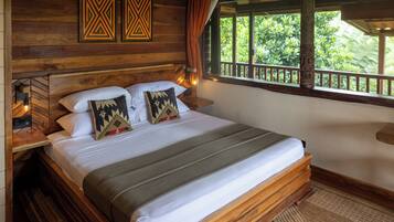 A wooden-framed bed with white linens and patterned pillows, a nightstand with a lamp, and a window with a view of greenery.
