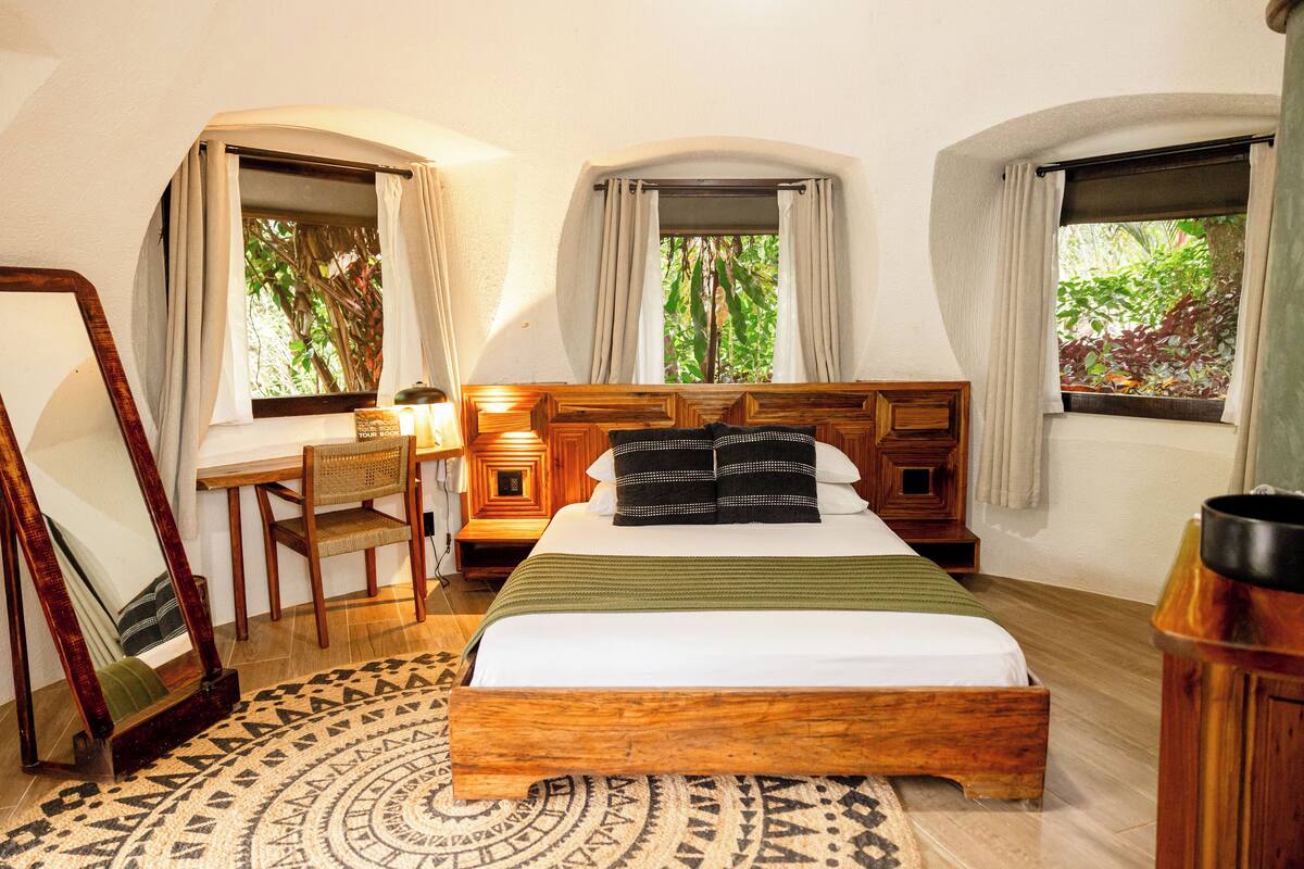 A bedroom with a wooden bed, a desk, and a chair, with windows offering views of greenery.