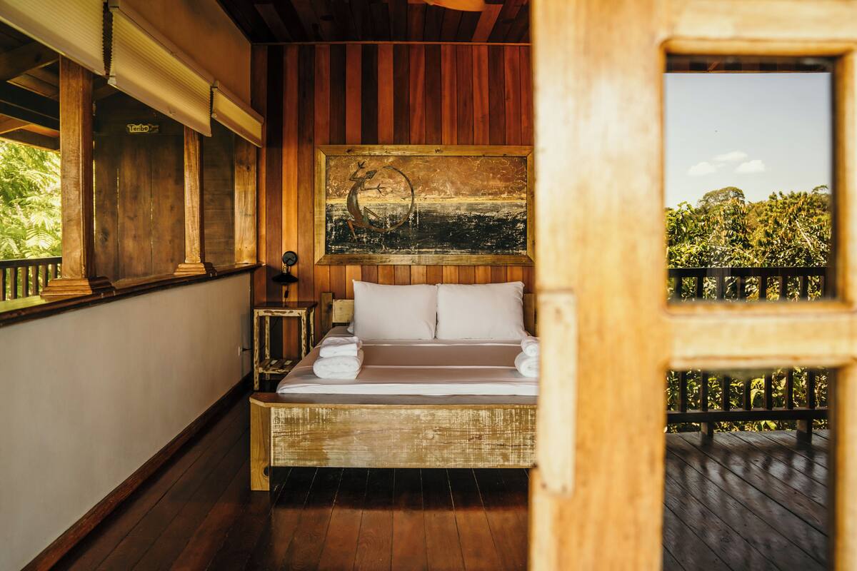 A wooden room with a bed, a painting, and a view of greenery.