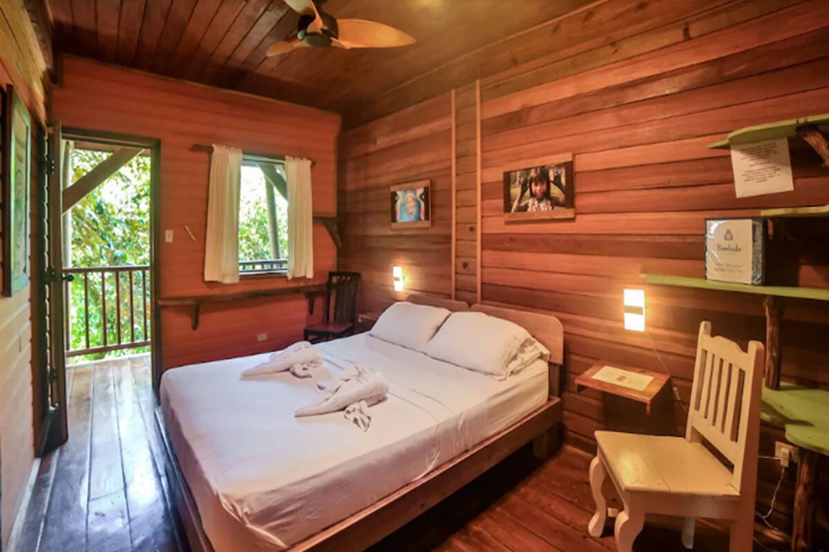 A wooden cabin room with a bed, a chair, a small table, and a balcony with a railing.