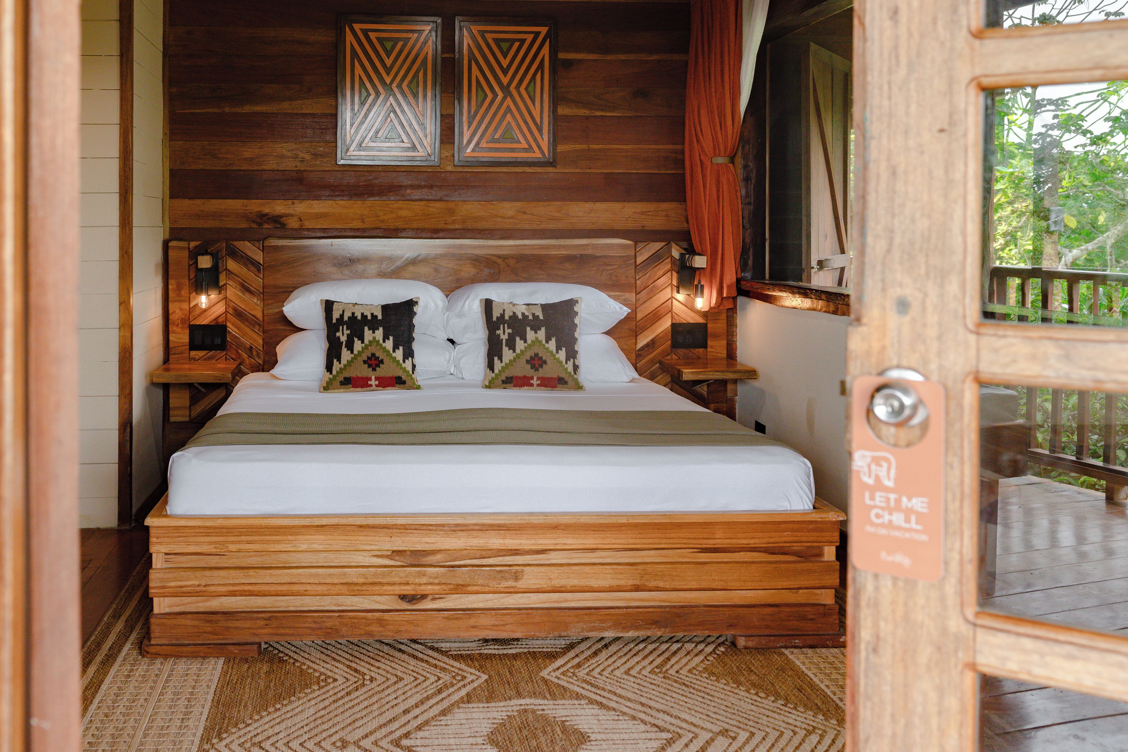 A wooden bed with two pillows, a nightstand with a lamp, and a herringbone-patterned wall.