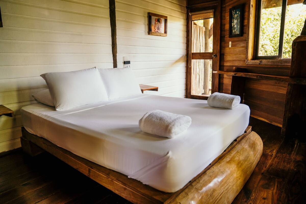 A wooden bed with white linens and two towels, a small bedside table, and a window with curtains.