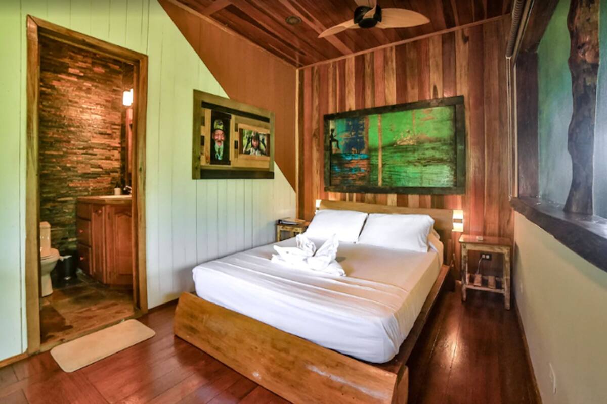 A bedroom with a wooden bed, a painting on the wall, and a wooden door leading to a bathroom.
