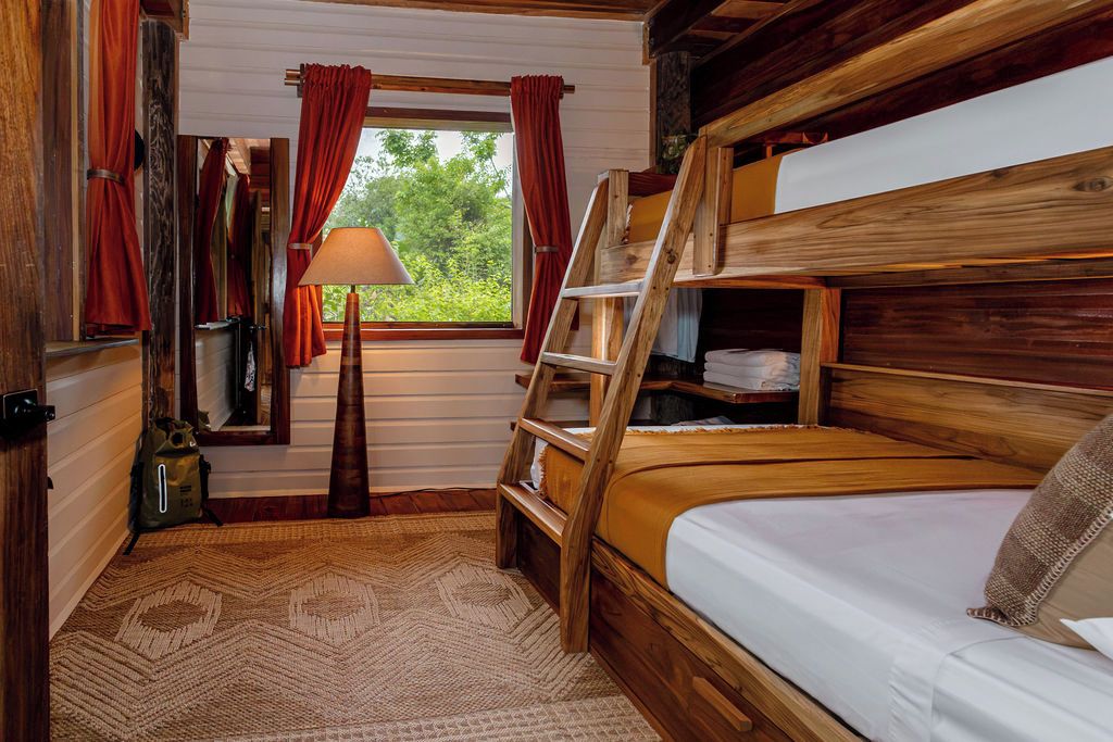 A cabin-style room with a bunk bed, a wooden ladder, a window with curtains, a lamp, and a rug.