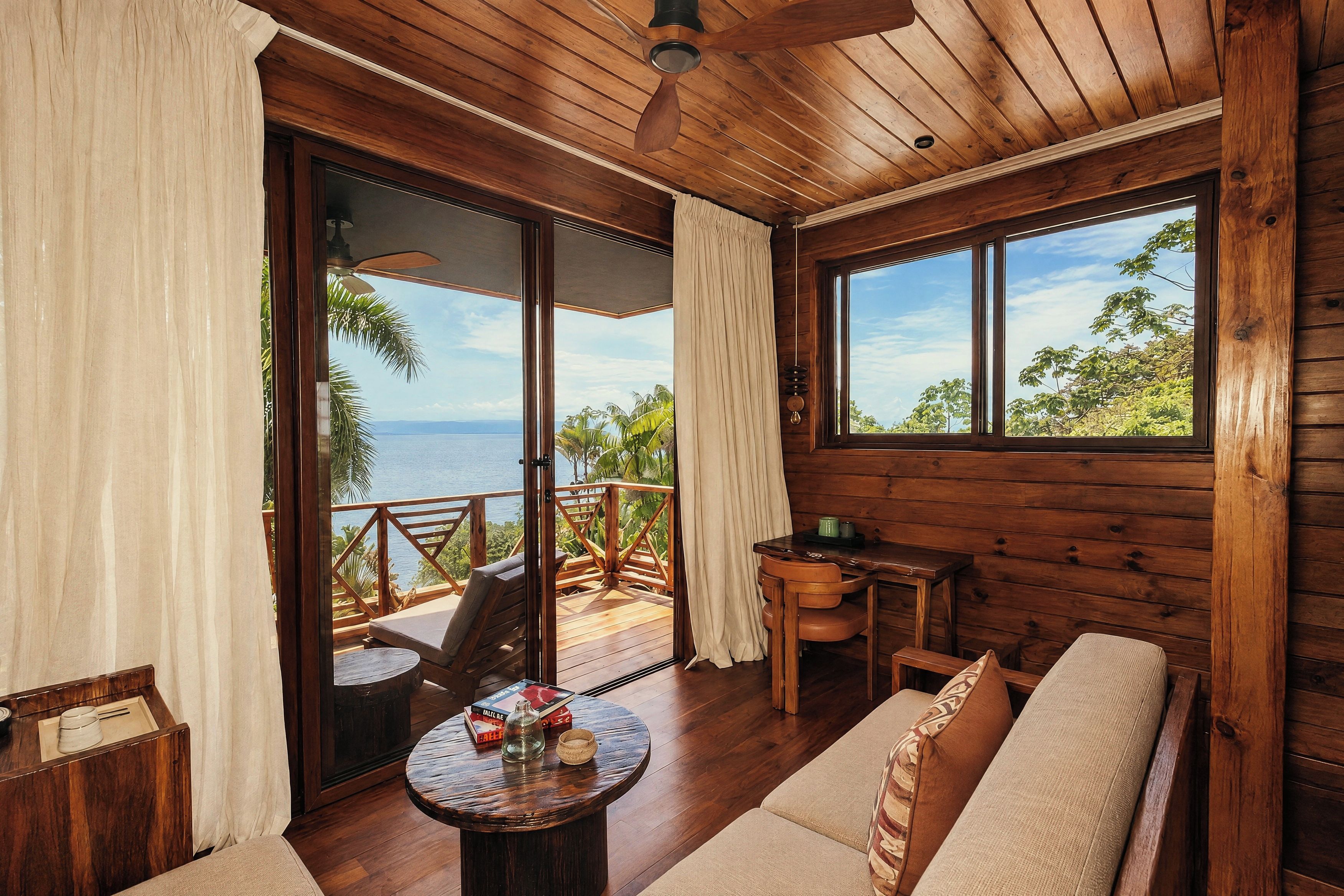 Interior de cabana de madeira com varanda, um sofá, uma mesa e vista para o oceano.