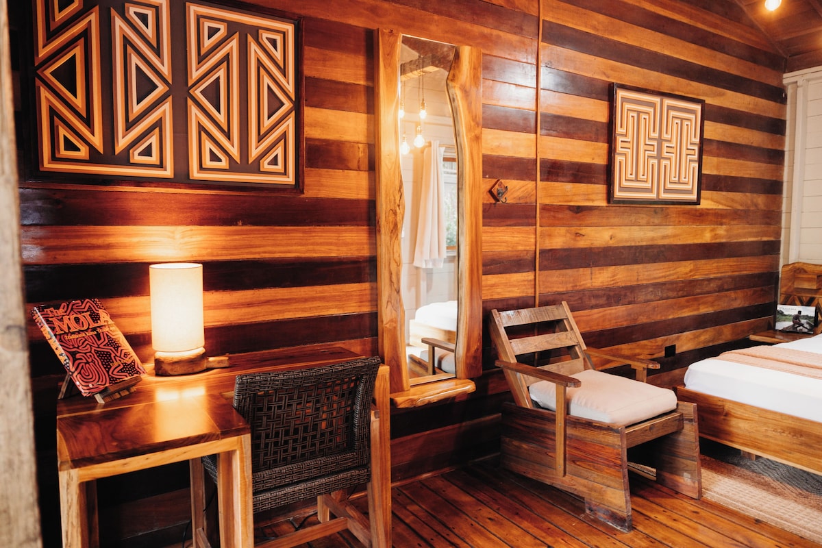A wooden cabin-style room with a bed, two chairs, a table with a lamp, and a mirror.